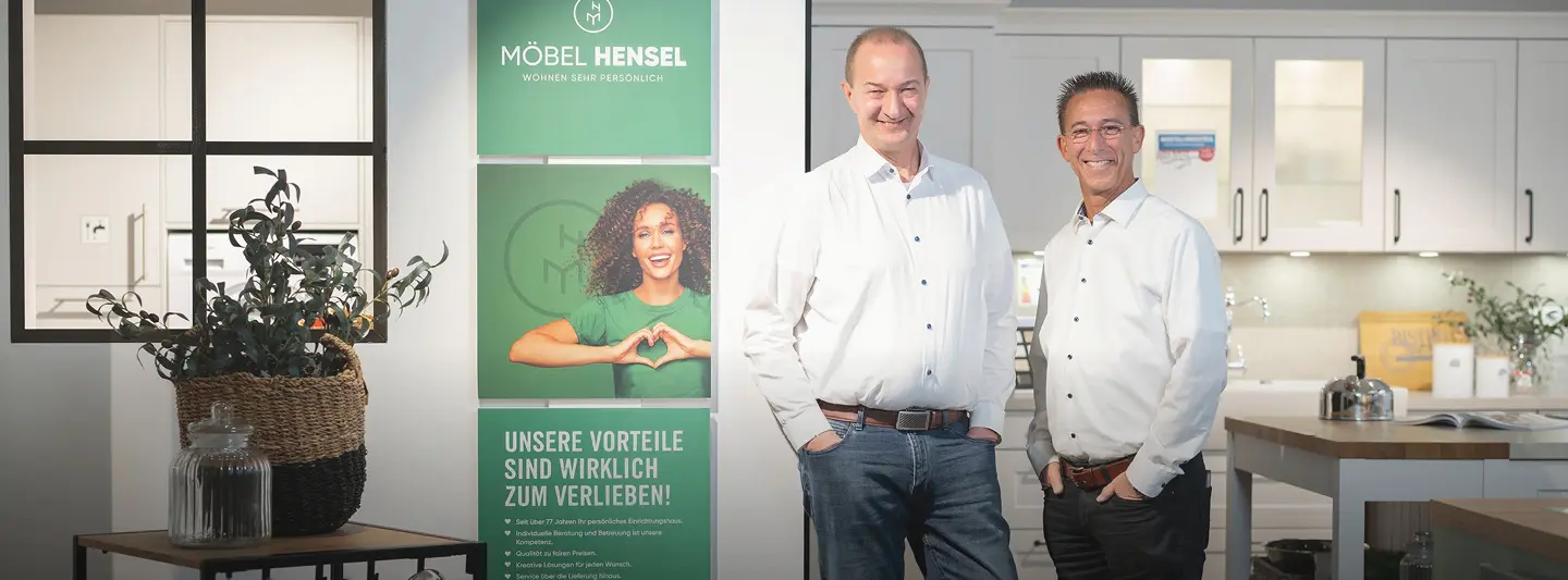 Zwei Männer stehen vor einer Küche, links grüne Poster mit Text Möbel Hensel und Unsere Vorteile sind wirklich zum Verlieben